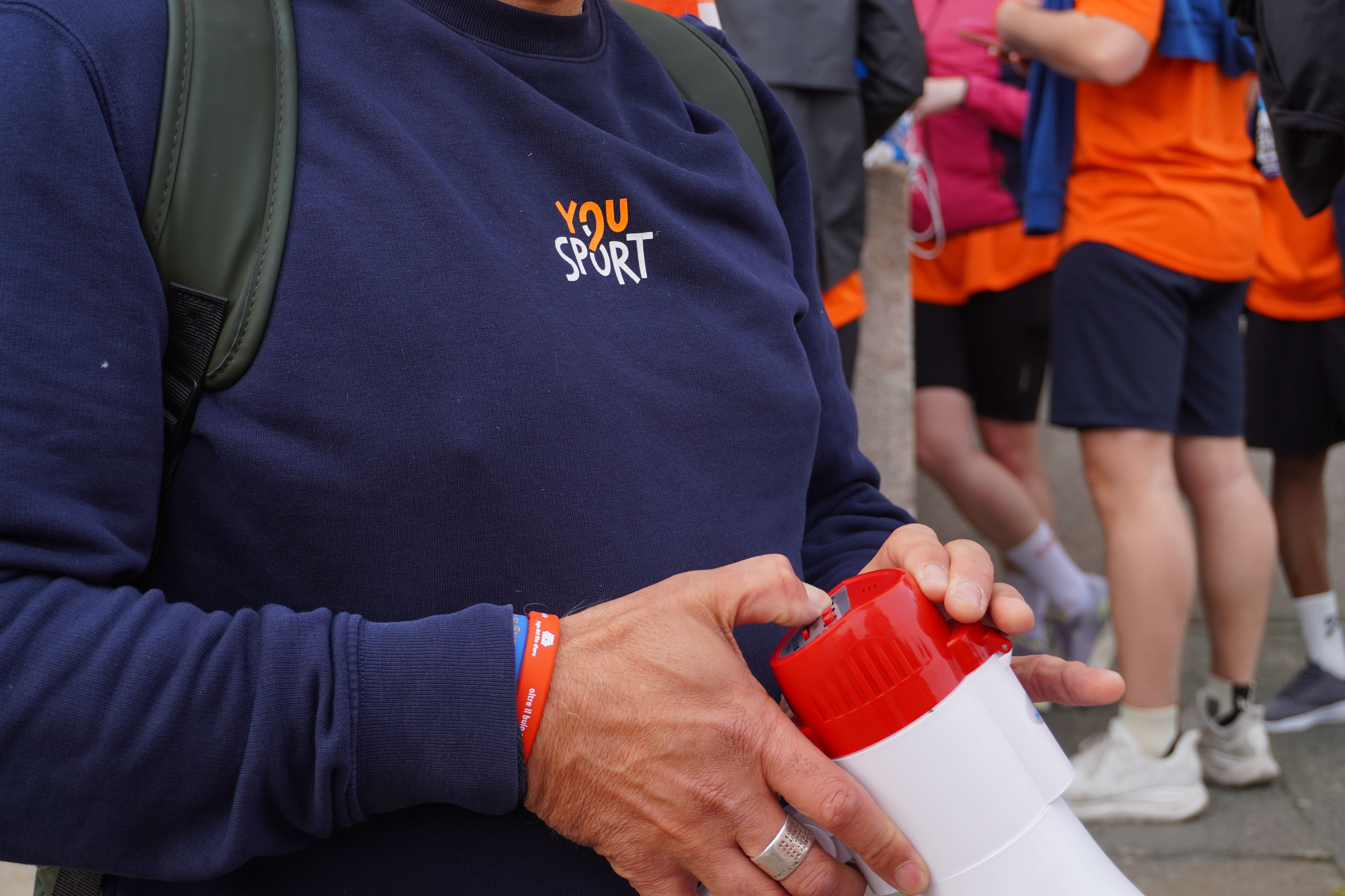 Due donne sorridenti con magliette arancioni etichettate 'You Sport' durante una maratona di staffetta, con volti truccati, circondate da altri corridori.