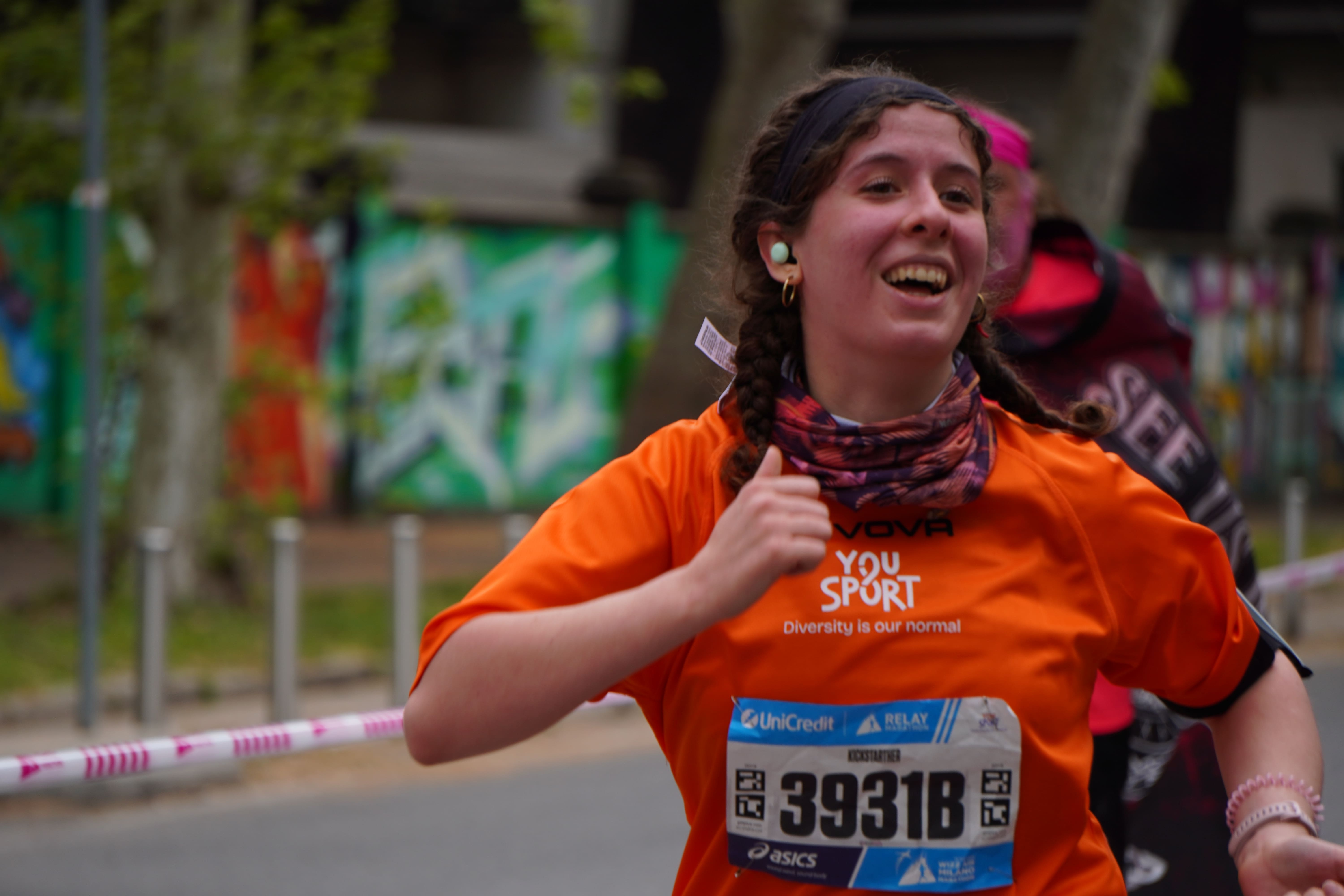 Gruppo di cinque donne con magliette arancioni YouSport e una bambina che soffia in una trombetta, tutte sorridenti dietro una cornice con scritta "Diversity is our normal" e disegni di palloni sportivi.