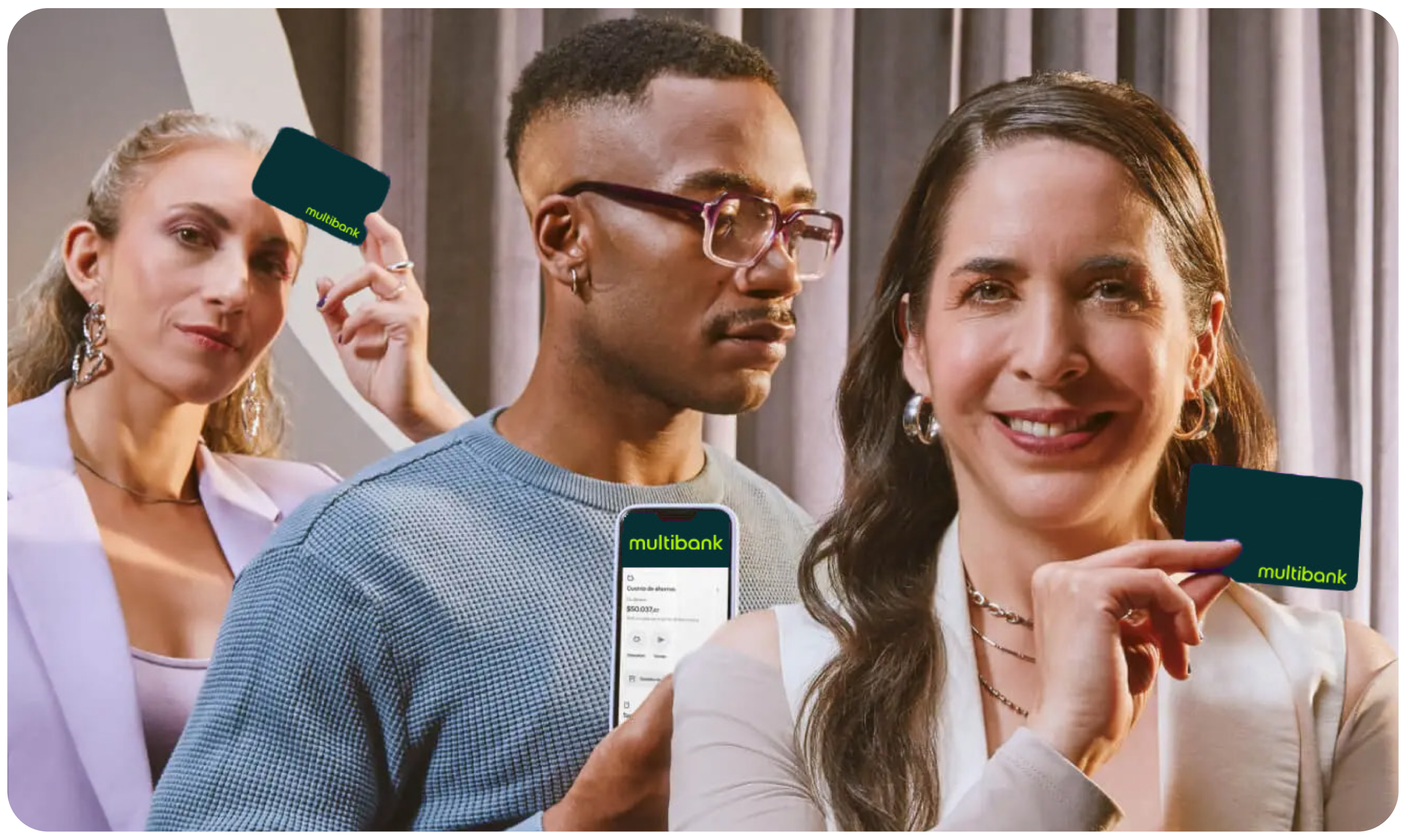 Three individuals smiling and holding Multibank cards in a modern setting with gray curtains. One shows a smartphone screen with a banking app.