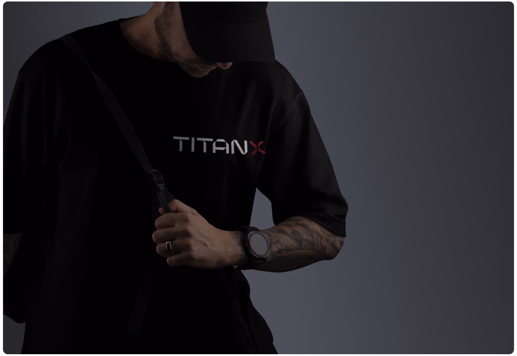 A person wearing a black "TitanX" t-shirt and cap in a dimly lit setting looks down, adjusting a strap. Their tattooed arm shows a watch. 