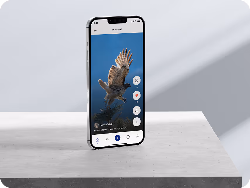 A smartphone stands on a gray surface displaying an app with a hawk in flight on a clear blue sky background. The interface shows engagement icons.
