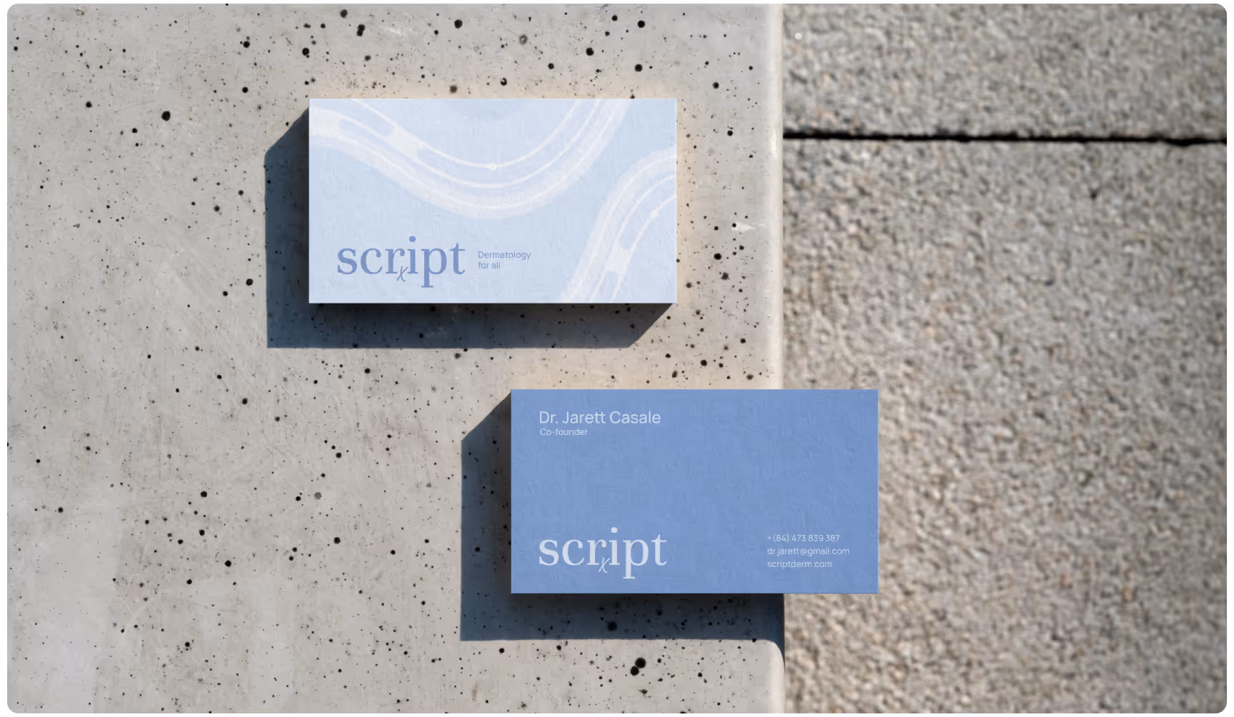 Two blue business cards on a textured concrete surface. One card shows abstract lines and "script" logo, the other lists a name and contact details. Minimalist and professional design.