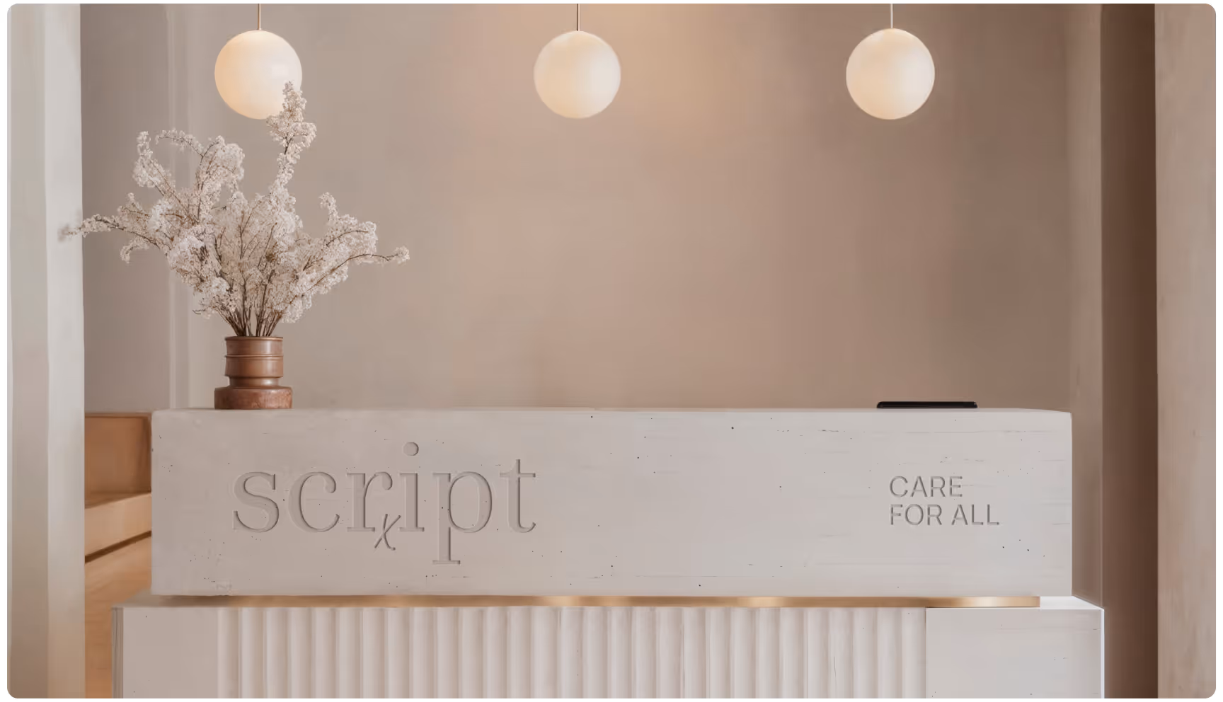 A modern reception desk with "script" and "CARE FOR ALL" engraved. Soft lighting, minimalist decor with white flowers in a vase, and three spherical lights above.