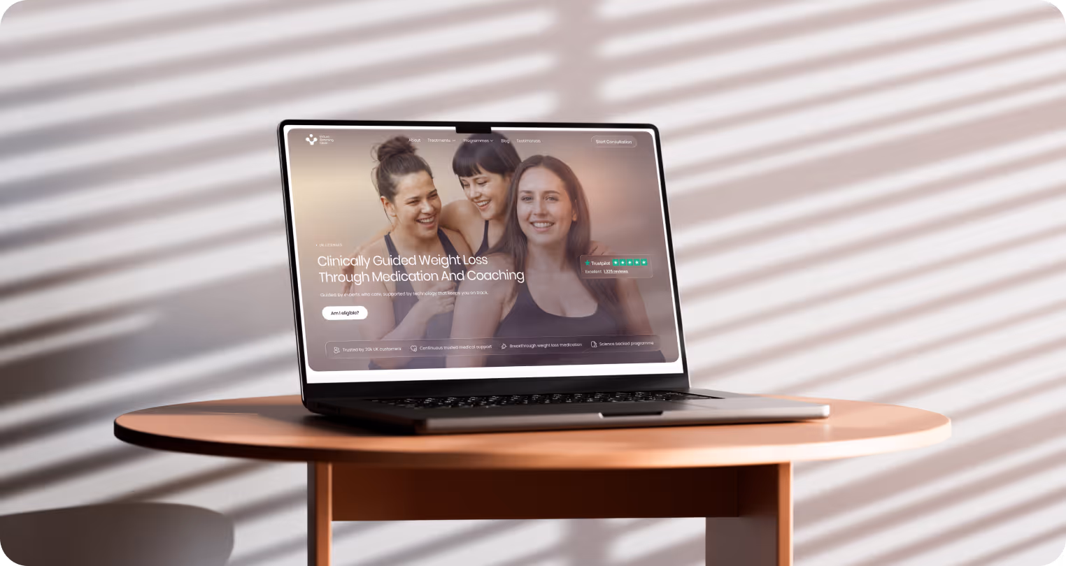 A laptop displaying a weight loss website on a round table, with three smiling women in the background. Sunlit blinds cast striped shadows, creating a warm tone.