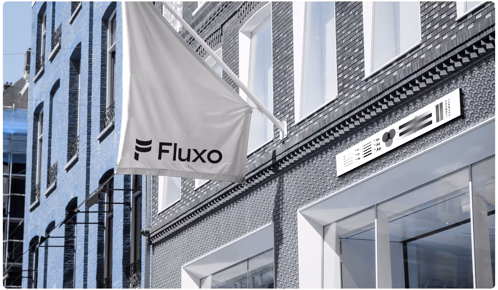 A white flag with the word "Fluxo" hangs outside a modern brick building with large windows and a monochrome sign above the entrance.
