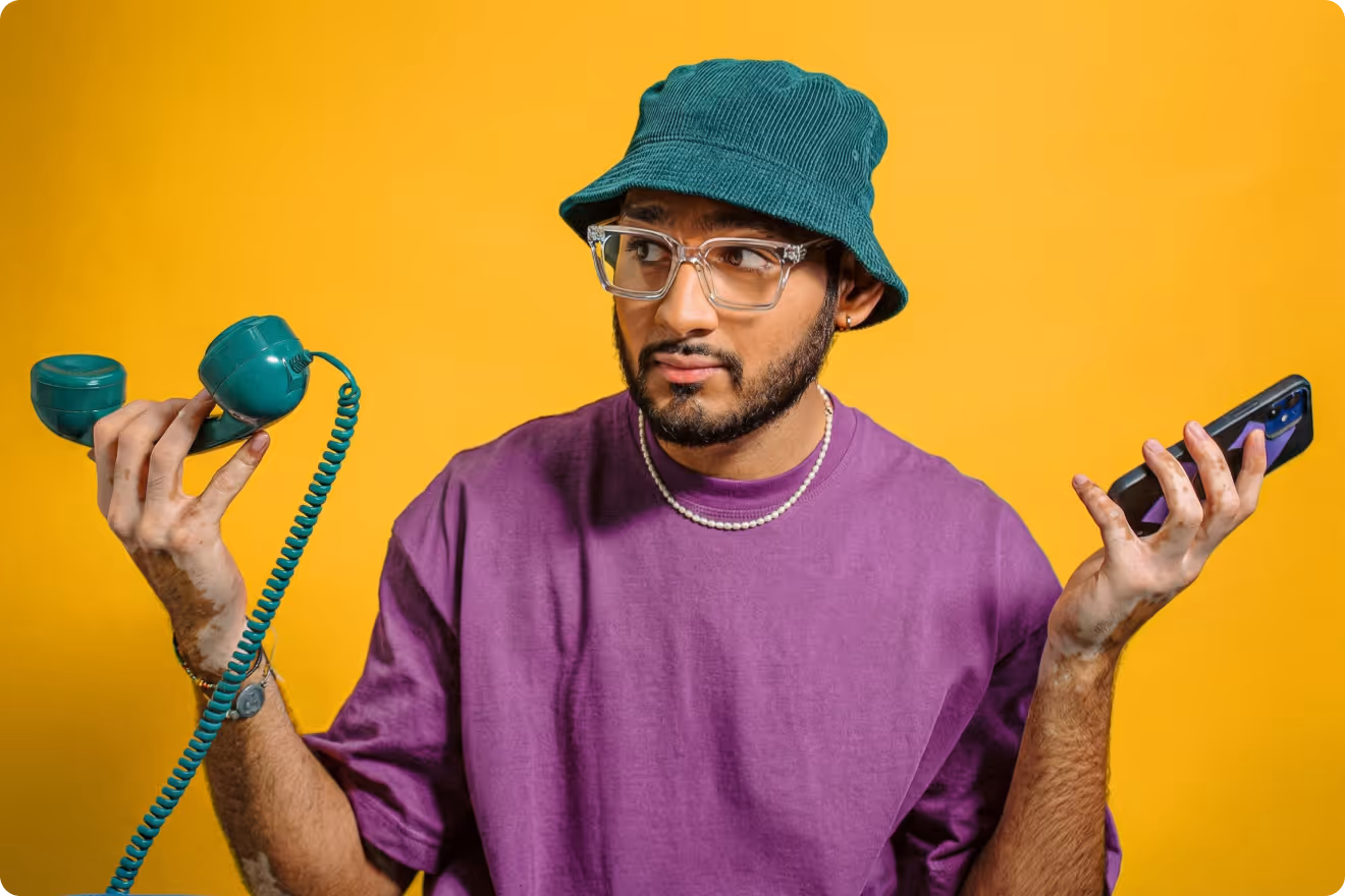 Person wearing a teal bucket hat, clear glasses, and a purple shirt holding a vintage corded telephone in one hand and a smartphone in the other against a bright yellow background, illustrating communication across generations and modern technology.