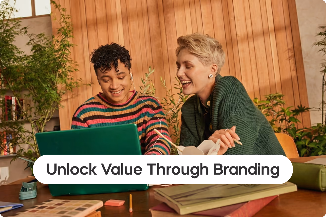 Two colleagues smiling and collaborating around a laptop in a warm, modern workspace, surrounded by design materials and plants, with the headline “Unlock Value Through Branding” displayed prominently across the bottom.