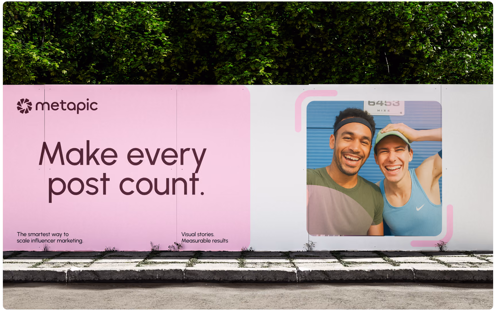 A wide street-level Metapic ad poster on a wall next to green foliage, featuring the slogan "Make every post count." alongside a photo of two smiling athletic men.