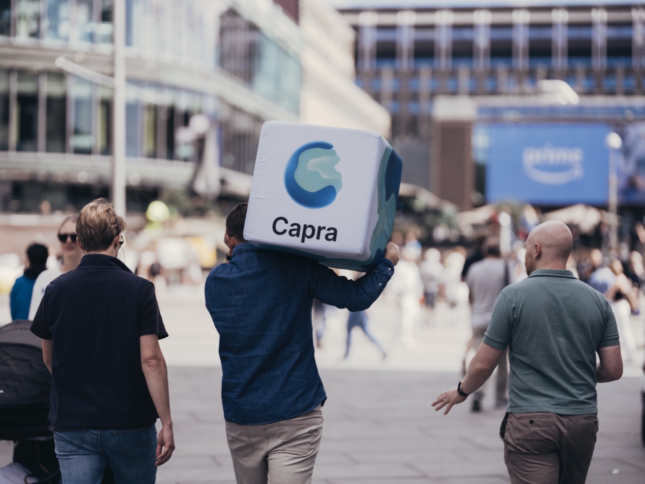 Tre menn går bortover en bygate, hvor en av dem bærer en stor kube med Capra-logo på skulderen.