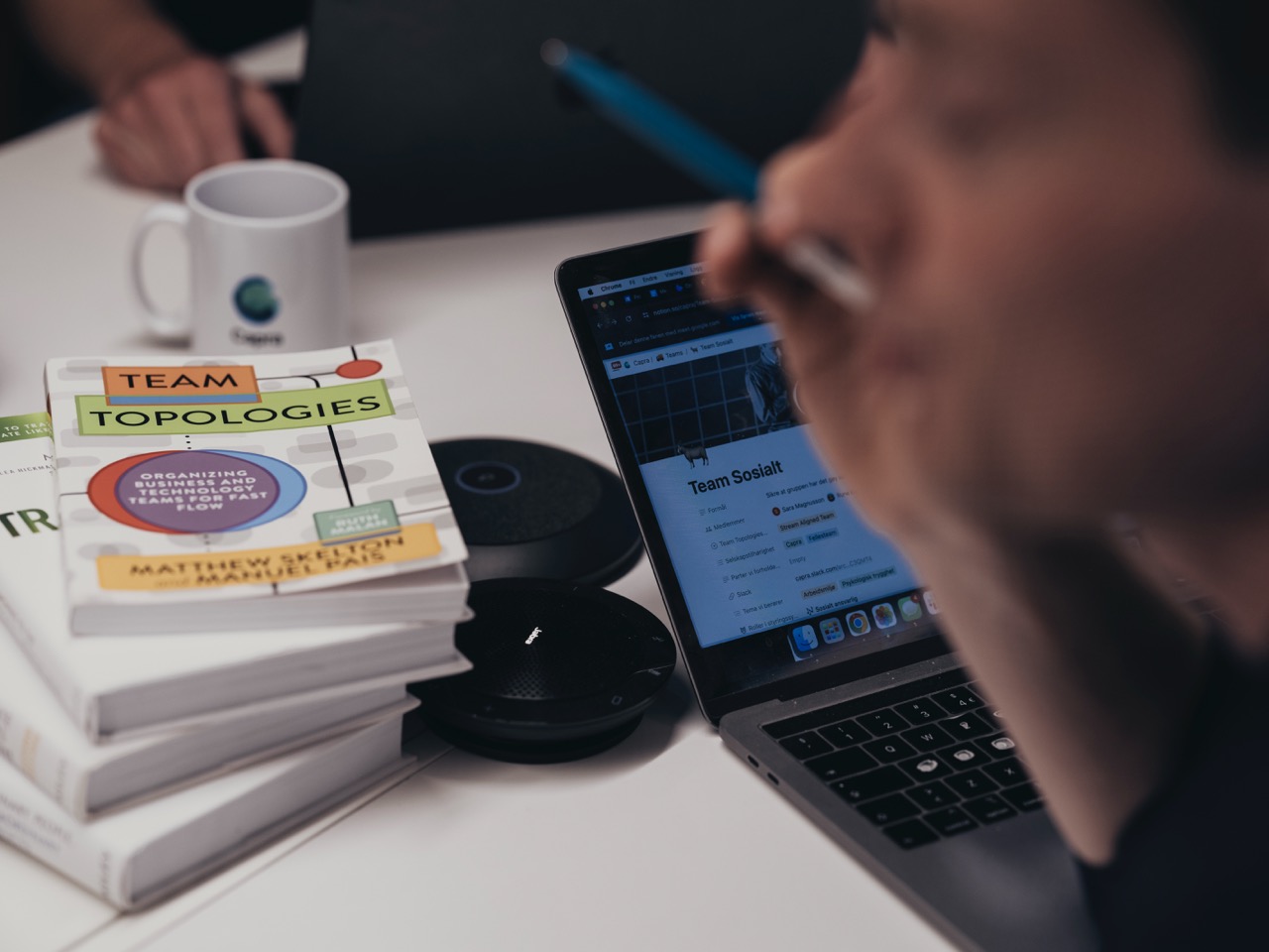 Nærbilde av hender ved en bærbar PC som viser en nettside for Team Sosialt, med en bokstabel og kaffekopp på et skrivebord.