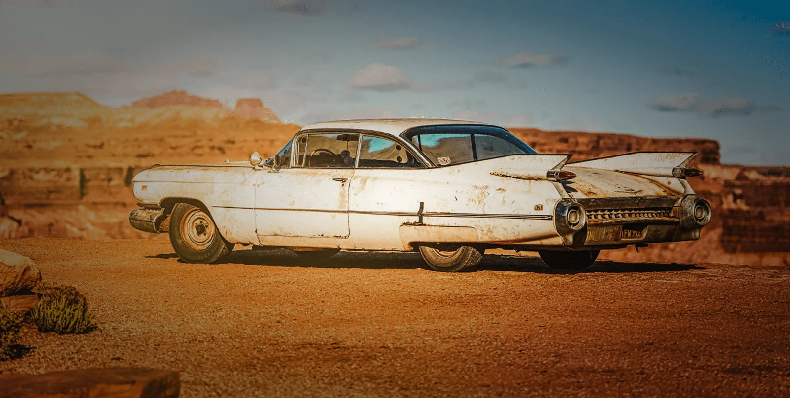 Rusty white vintage Cadillac parked on a desert overlook with red rock canyons in the background.