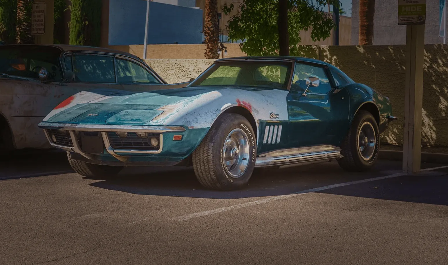 Worn blue classic Corvette parked in a shaded lot beside another vintage car.