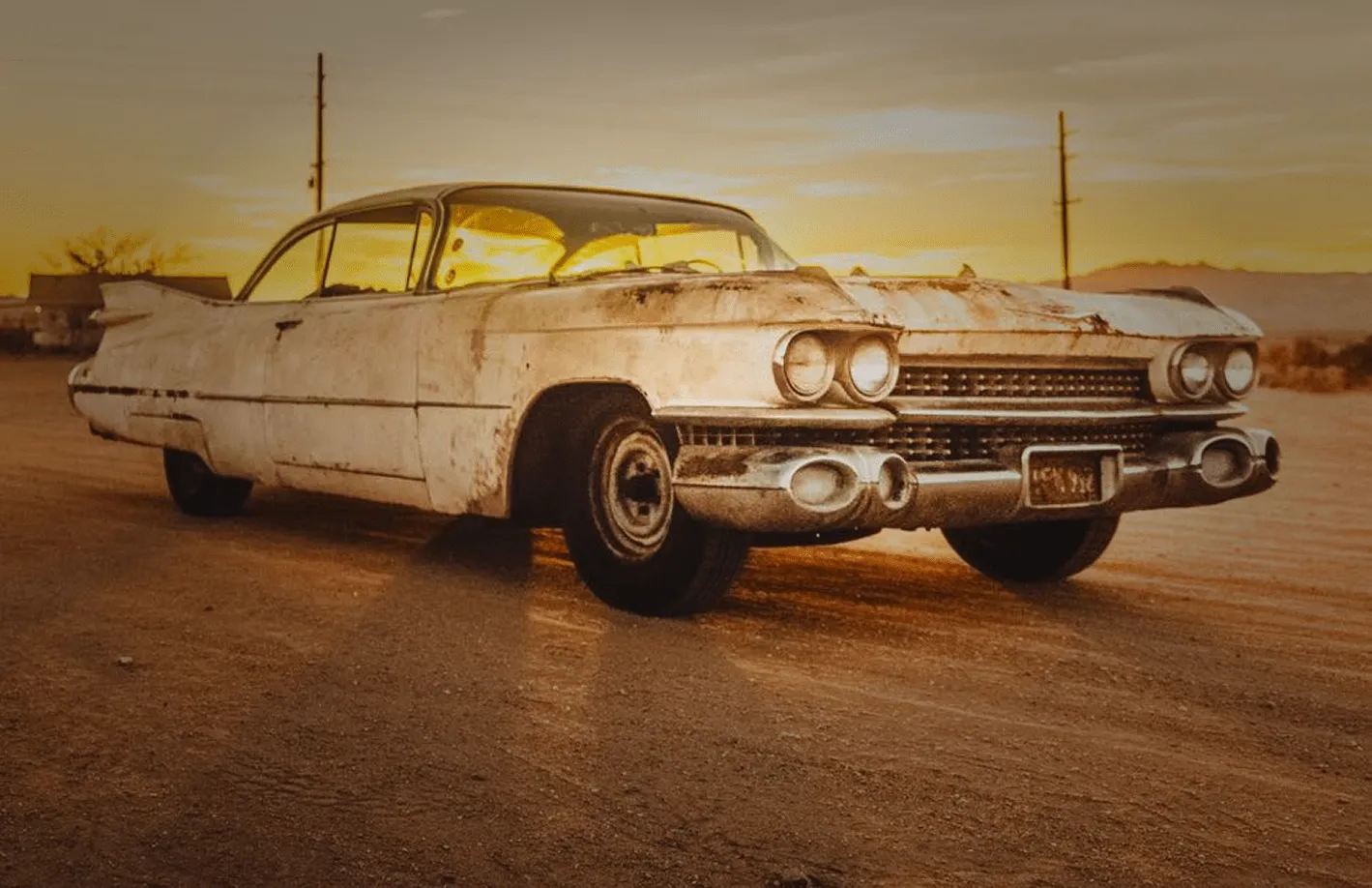 Rusty vintage Cadillac on a dirt road at sunset with golden light reflecting off the car.