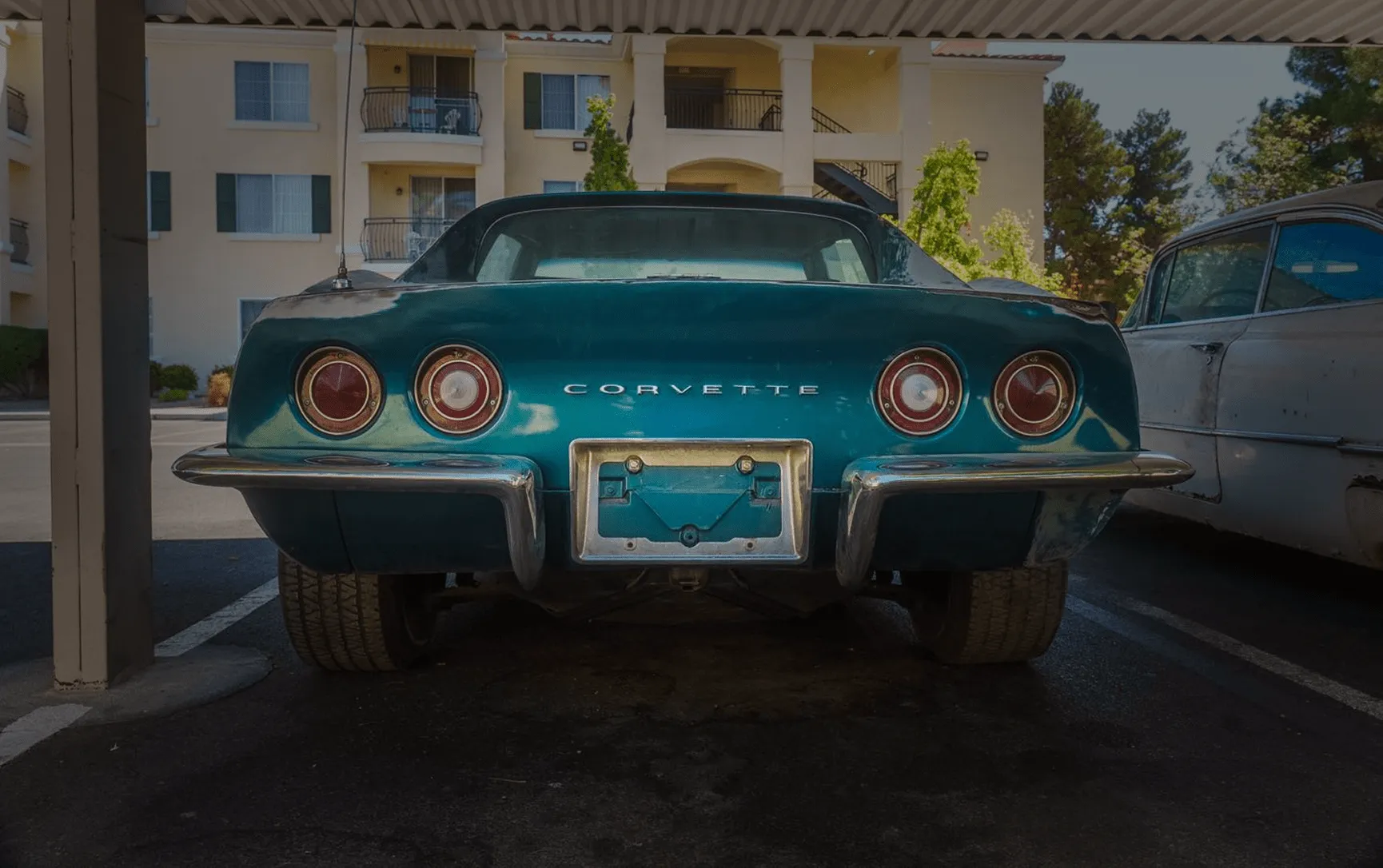 Rear view of a teal vintage Corvette parked under a carport beside another old car.