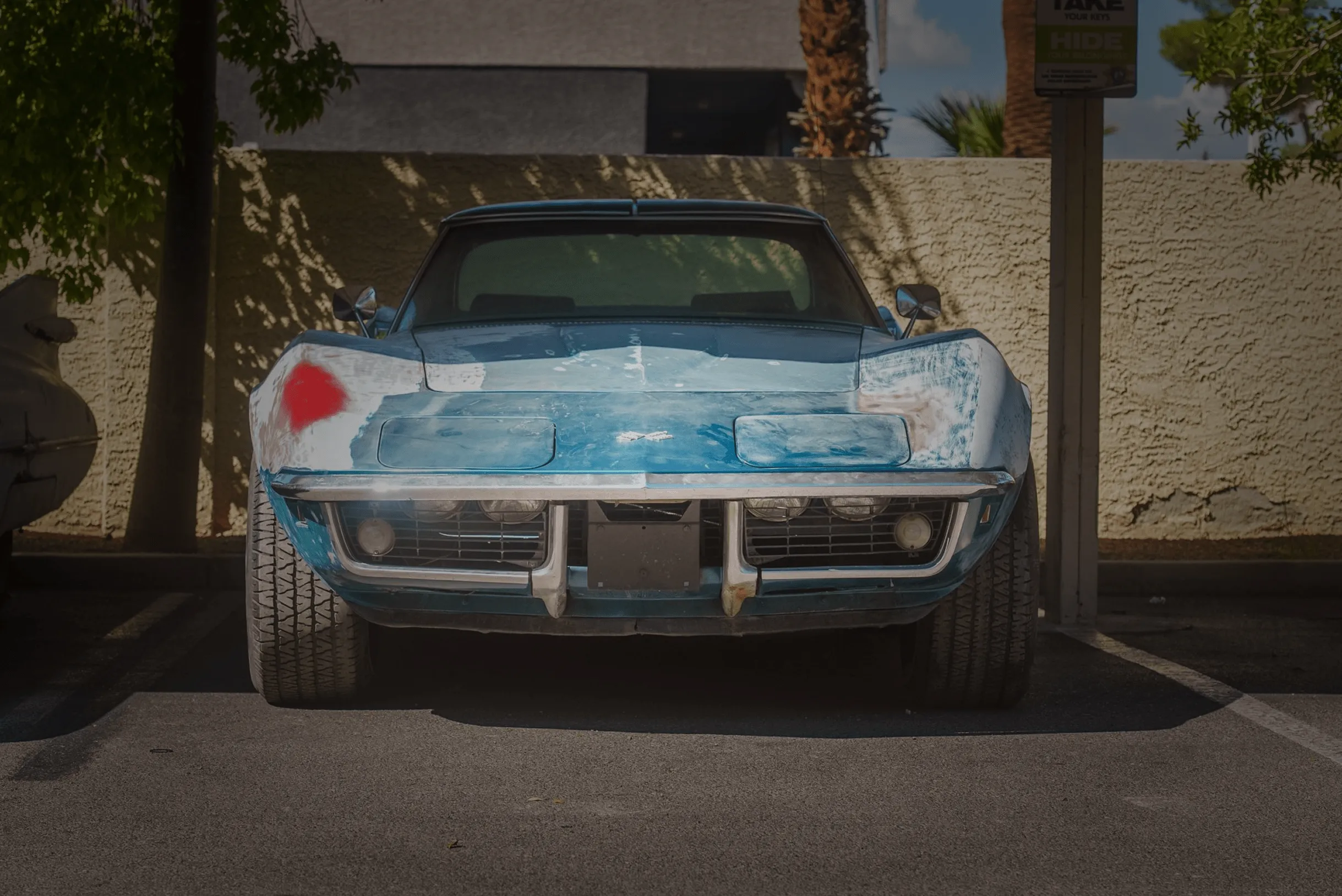 Front view of a weathered blue Corvette with faded paint and a red primer spot, parked in a sunny lot.