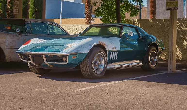 Worn blue classic Corvette parked in a shaded lot beside another vintage car.