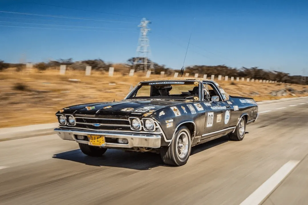 Black vintage Chevrolet El Camino with racing decals driving on a desert highway.