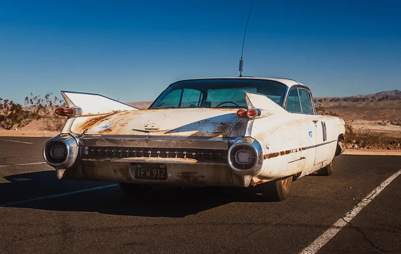 Rear view of a rusty white vintage Cadillac parked on an empty desert road under a clear blue sky.