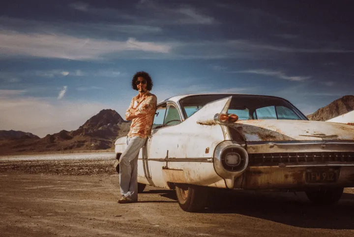 Person leaning against a rusty vintage Cadillac on a desert road with mountains in the background.