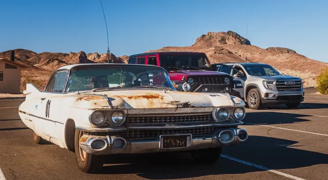 Rusty white vintage Cadillac parked beside modern SUVs in a desert lot with rocky hills in the background.
