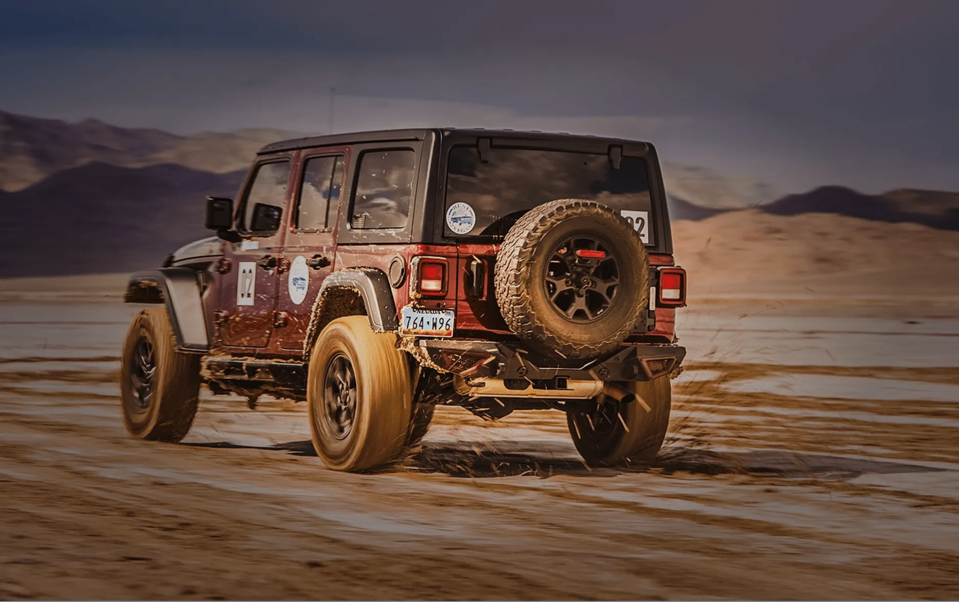 Rear view of an off-road SUV splashing through mud on a desert plain with mountains in the distance.