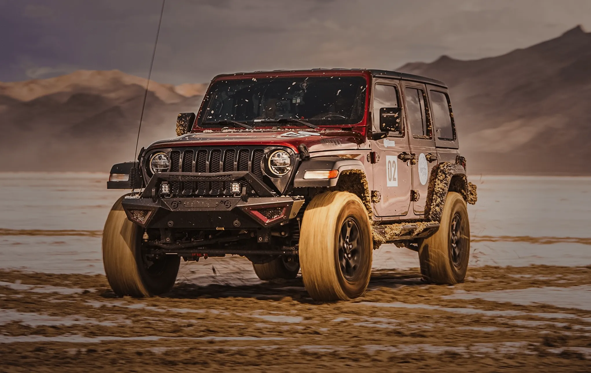 Off-road SUV kicking up mud while driving across a rugged desert landscape with mountains in the background.