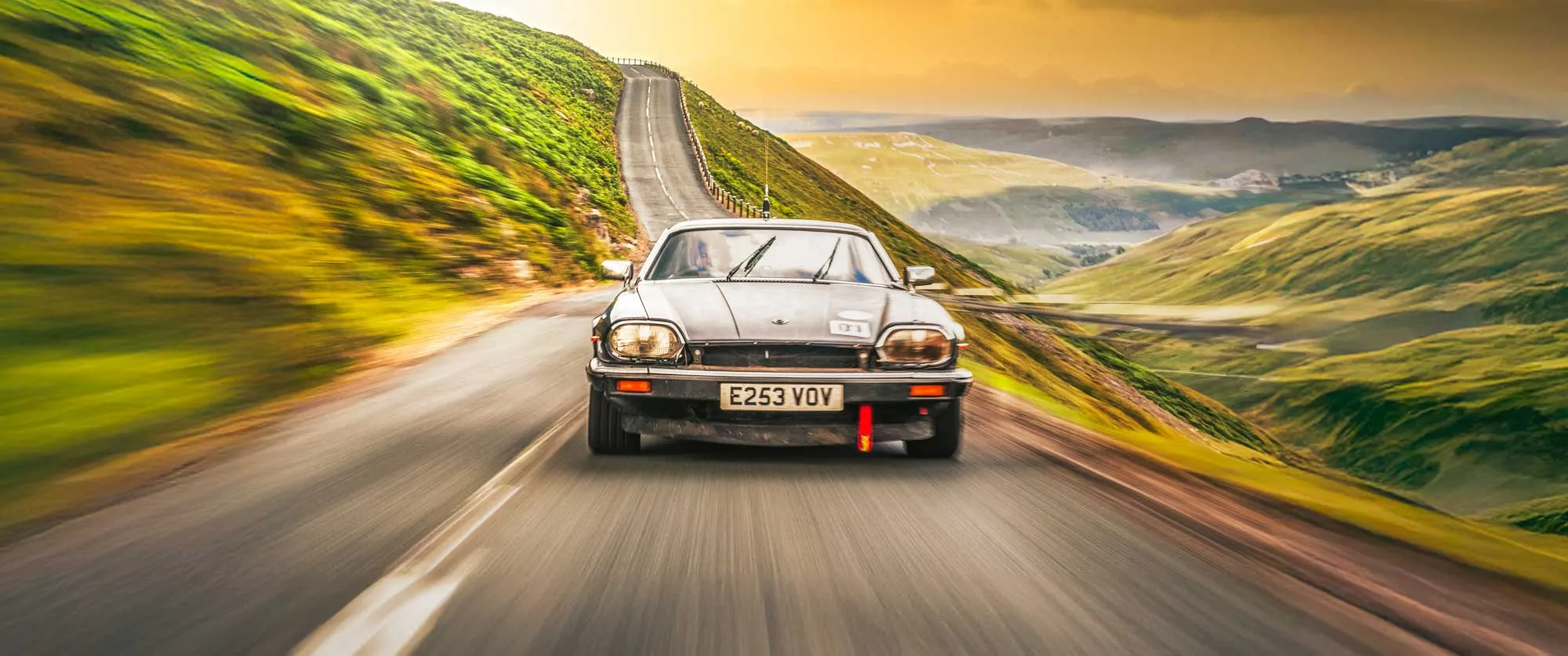A vintage black sports car driving at speed on a winding mountain road. The road curves uphill through lush green hills under a dramatic golden sunset sky. A motion blur effect emphasizes the car's movement and speed.