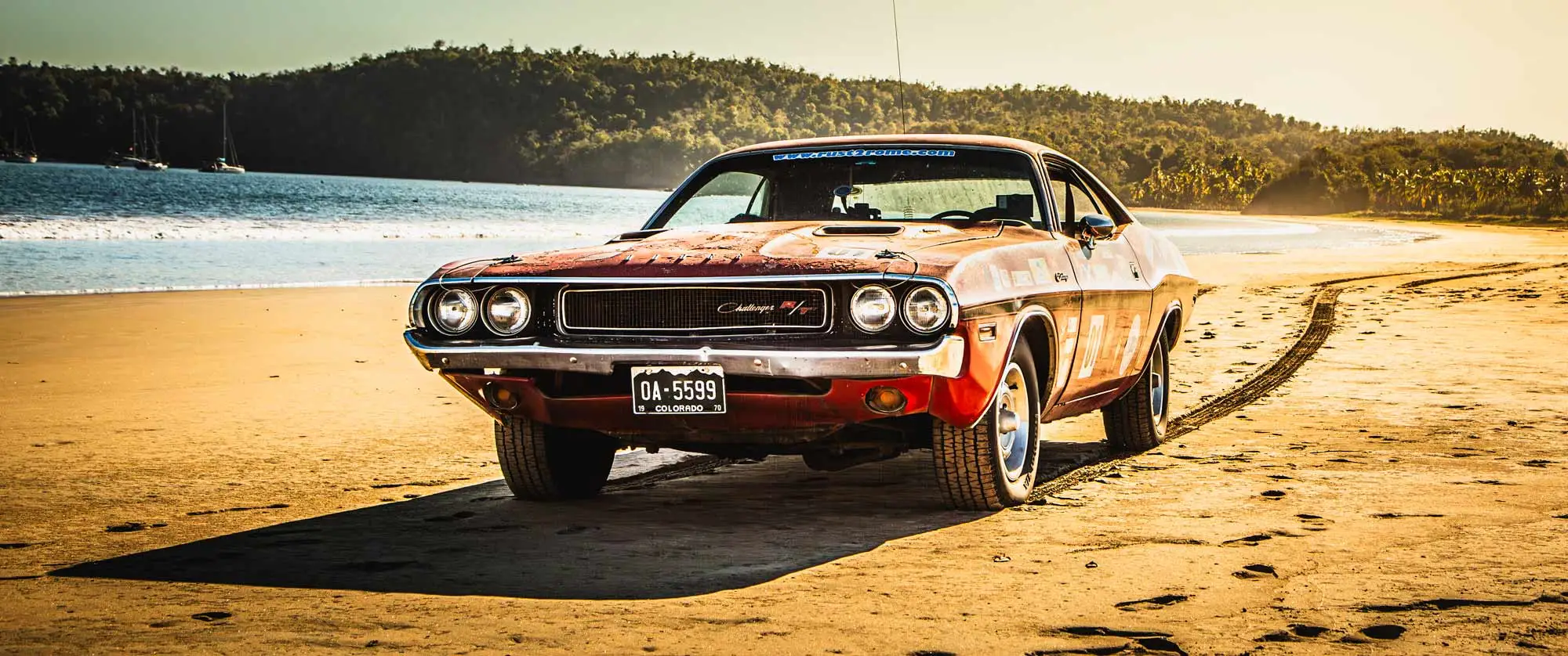 A classic muscle car parked on a sandy beach in Mexico at sunset, with tire tracks behind it and the ocean and forested hills in the background.