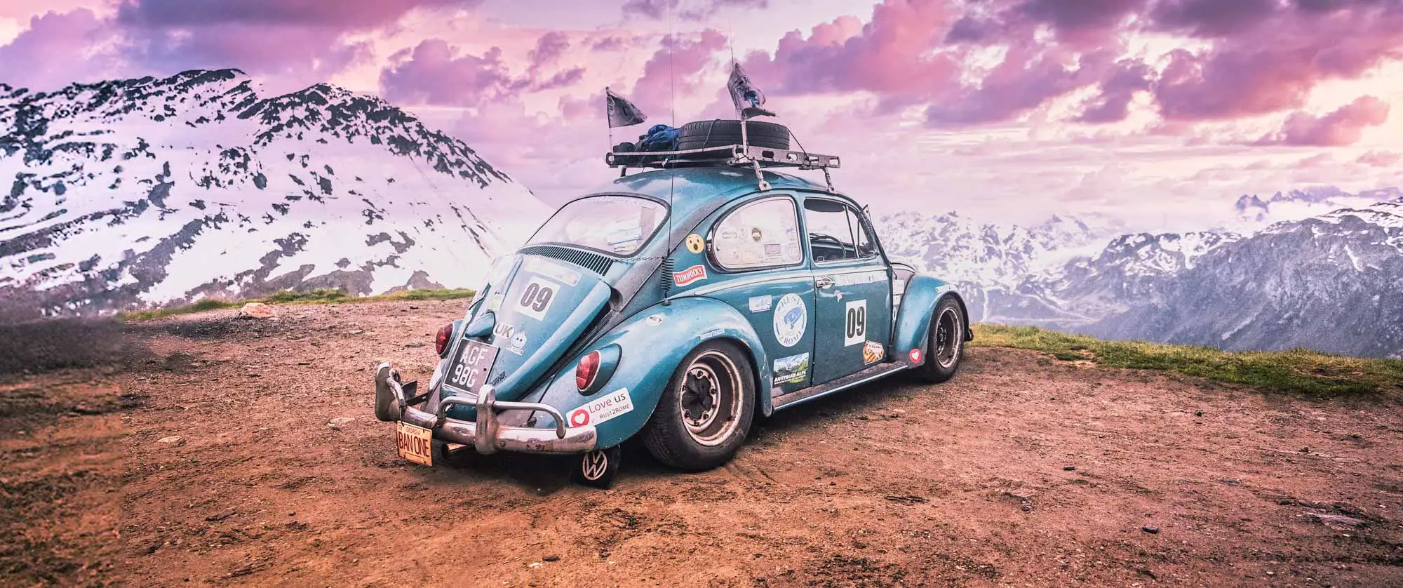 A vintage blue Volkswagen Beetle rally car parked on a mountain overlook, with snow-covered peaks and a dramatic pink and purple sky in the background.