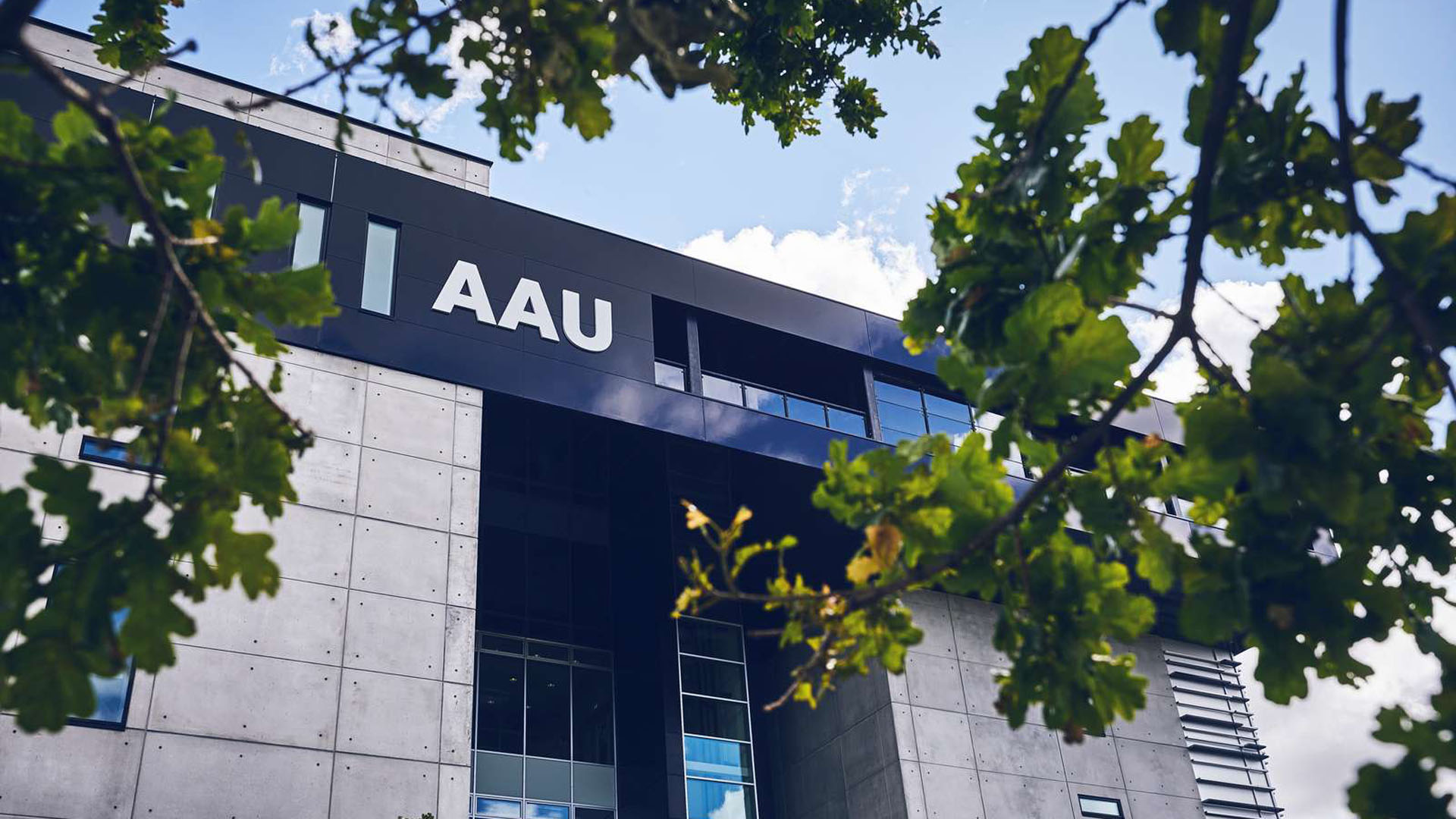 Aalborg Universitet København campus set udefra med moderne bygninger, glasfacade og universitetslogo. Efterårsfarvede træer står foran bygningen.