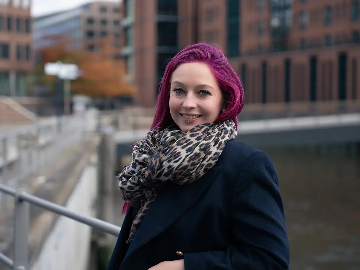 Porträtfoto von Cassandra Schwerter, Gründerin von Sent4Souls - CSG Projects, mit pink-violetten Haaren, dunkelblauem Mantel und Leopardenmuster-Schal. Sie steht draußen vor moderner Architektur an einem Wasserkanal und lächelt in die Kamera.