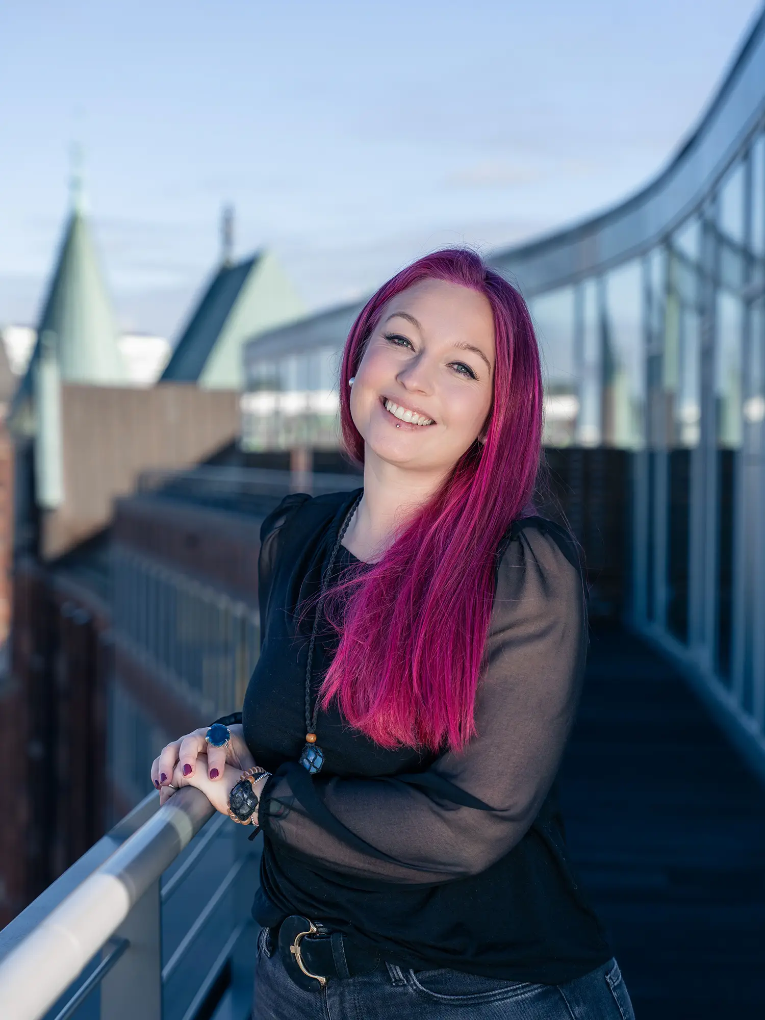 „Porträtfoto von Cassandra Schwerter, Gründerin von Sent4Souls - CSG Projects, mit pink-violetten Haaren und schwarzer Bluse. Sie steht auf einer Dachterrasse vor moderner Architektur, lehnt sich lächelnd an ein Geländer.