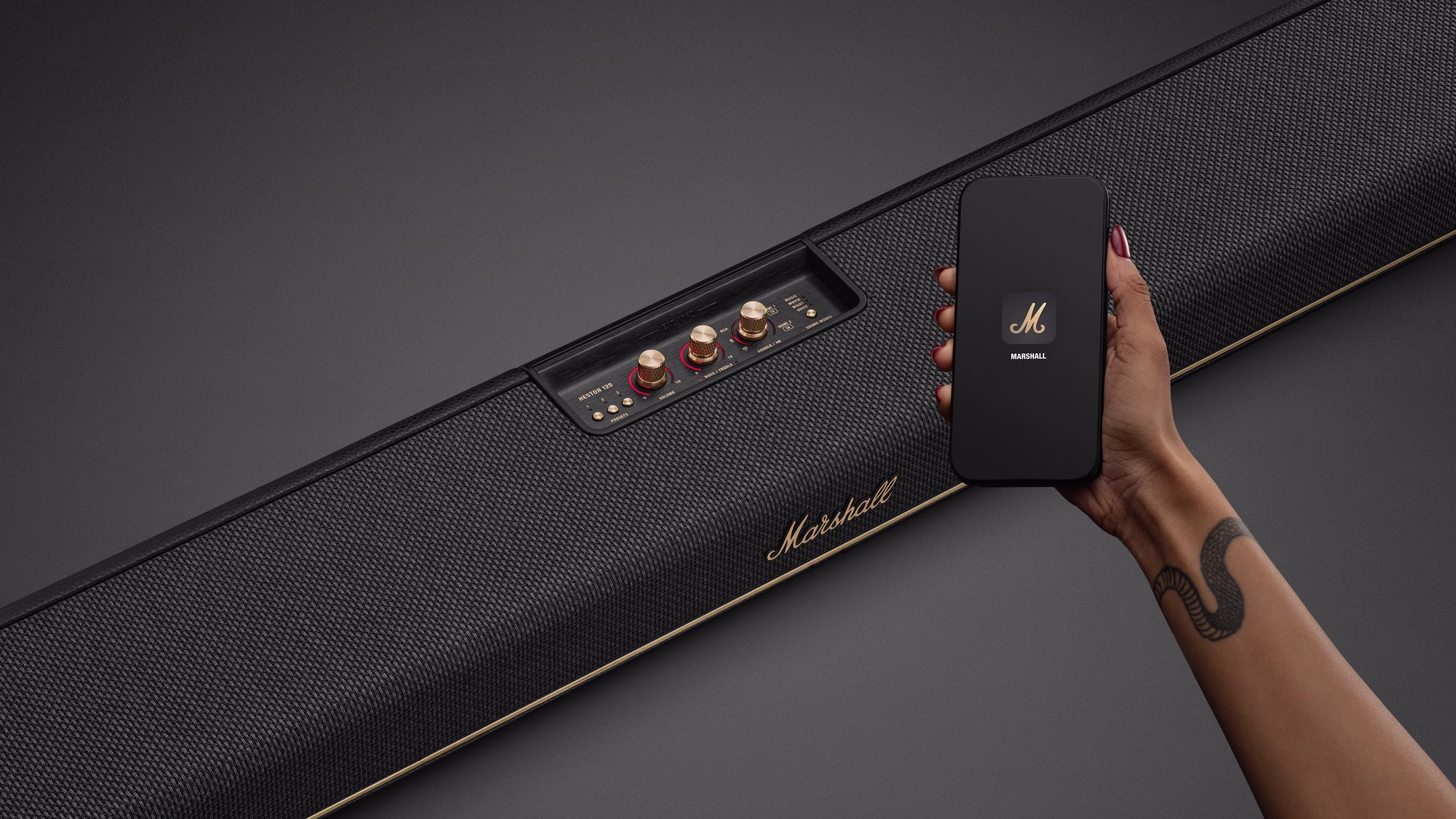 A hand holding a phone with the Marshall app symbol in front of a Heston 120 soundbar