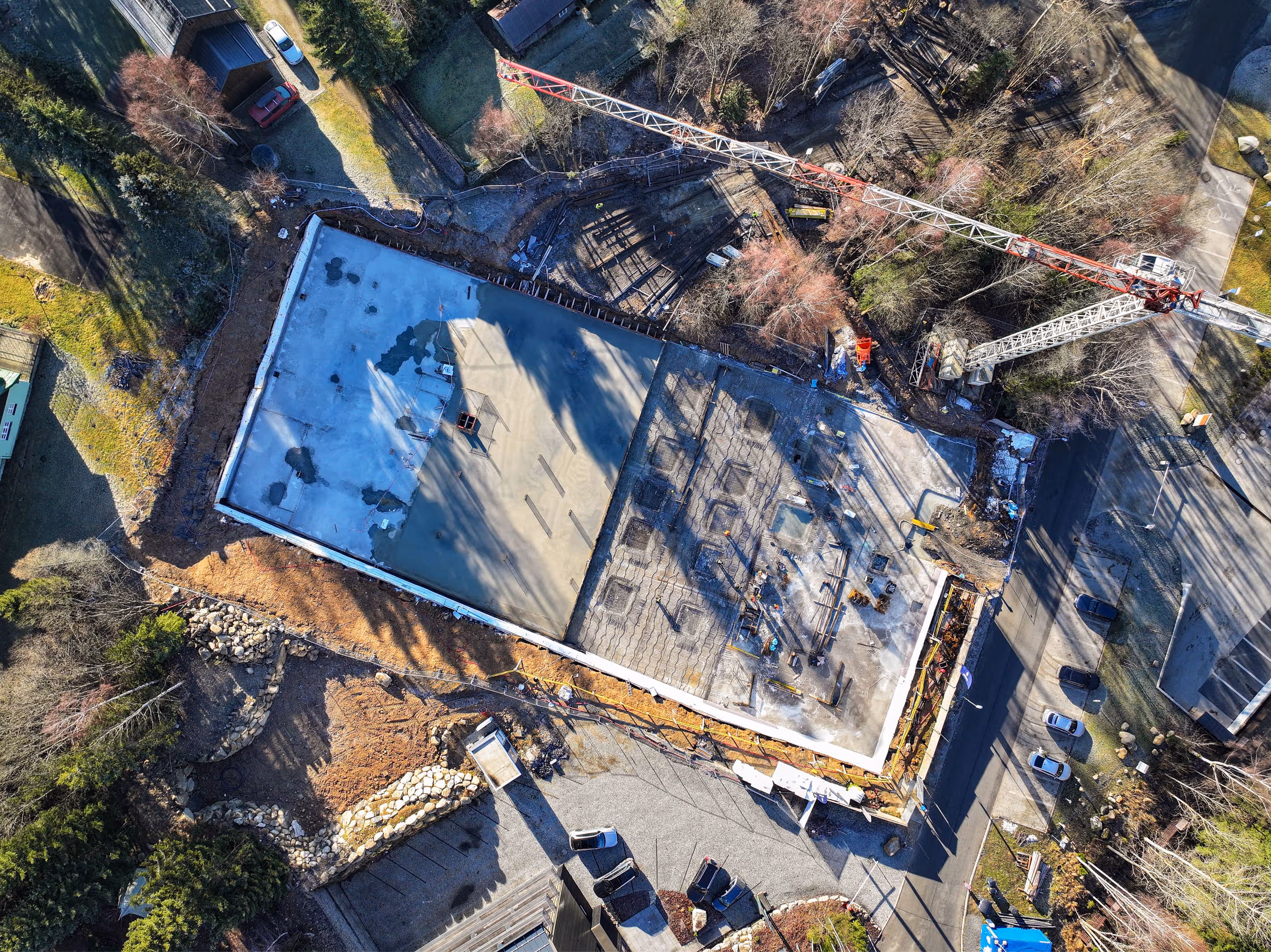 Letecký pohled na staveniště s betonovou základovou deskou, jeřábem a zaparkovanými auty v okolí.
