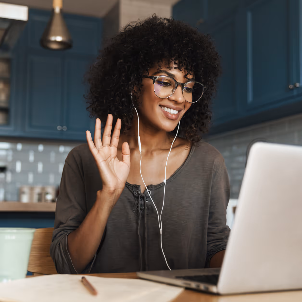 Smiling woman with curly hair having an online video call on a laptop, wearing earphones in a modern kitchen.