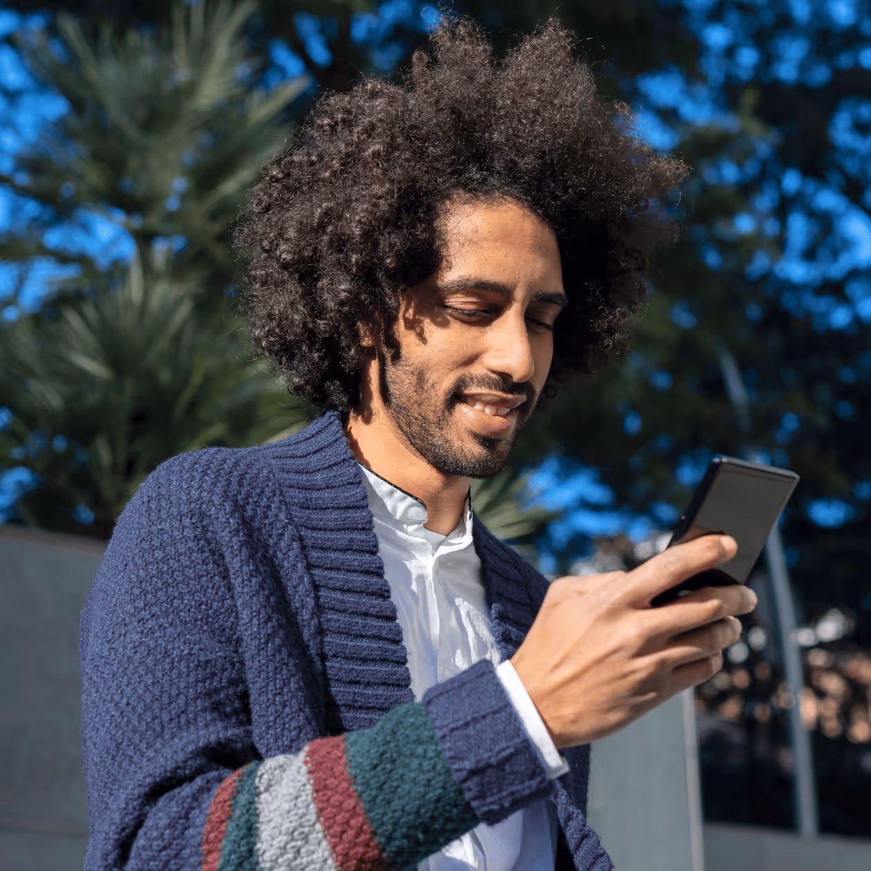 A young man with dark, curly hair and a beard smiles while looking at his smartphone, appearing engaged in a conversation or interaction.