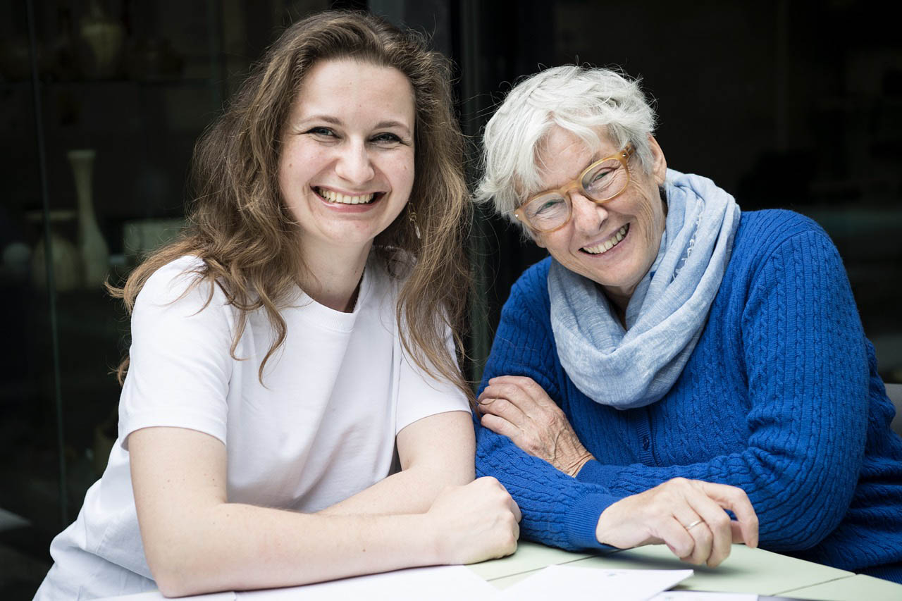 Une jeune femme avec de longs cheveux bruns sourit de toutes ses dents avec sa mentor à côté d'elle qui sourti également. La mentor est une femme âgée avec des cheveux blancs et des lunettes.