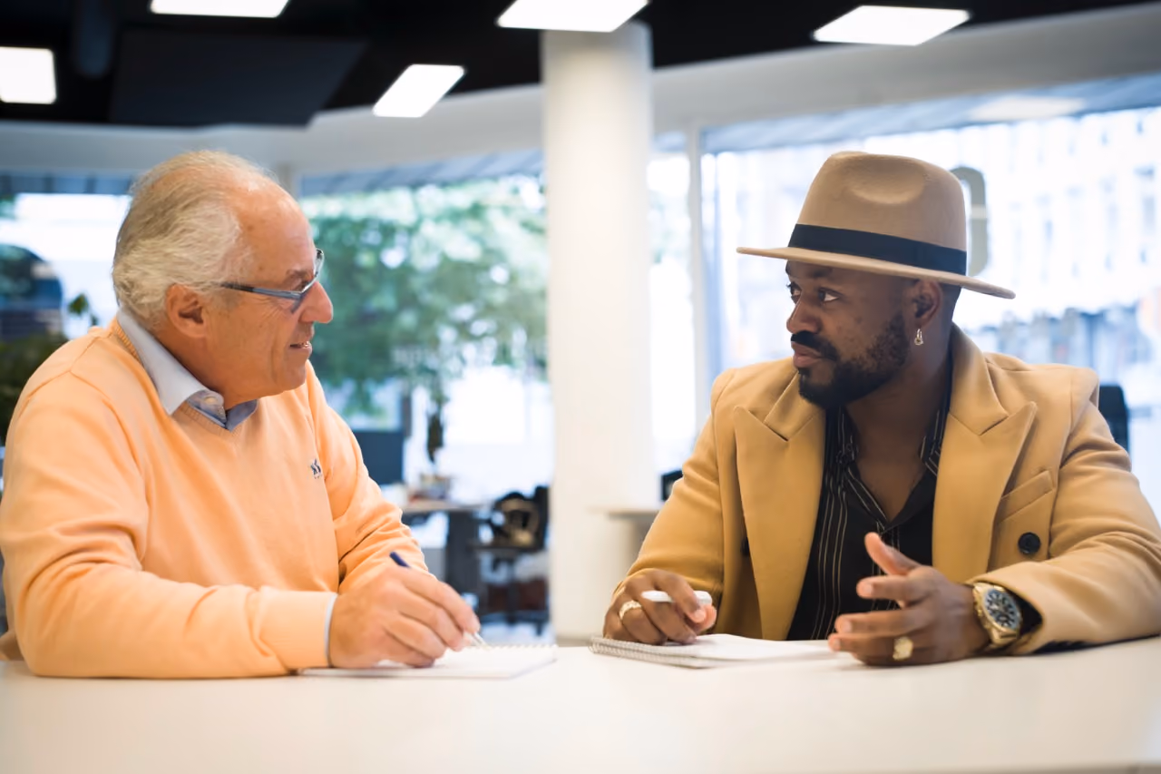 two people are sitting at a table talking. The first is a man over 60 with a salmon sweater, the second a black man with a beige Panama hat.