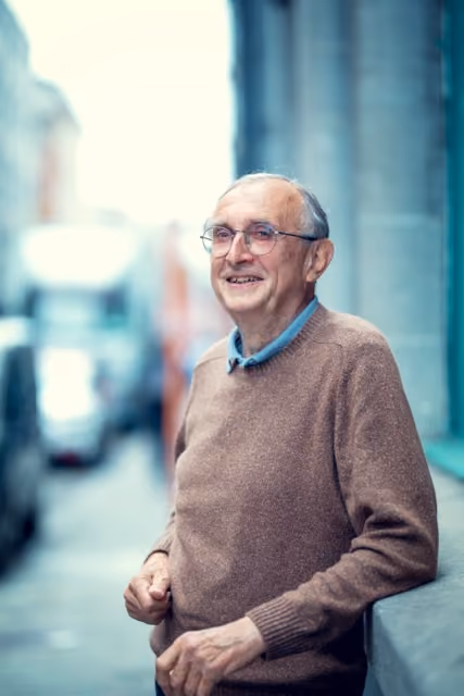 Portait d'un homme de plus de 70 ans avec un pull brun qu sourit en regardant le photographe