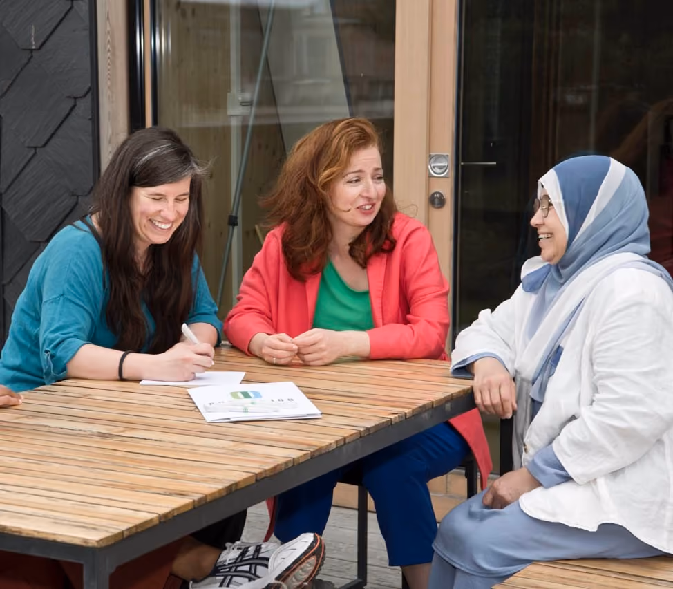 three women are talking around a wooden table on a terrace. The first is smiling while writing on a sheet, the second is talking to the third who is wearing a veil.