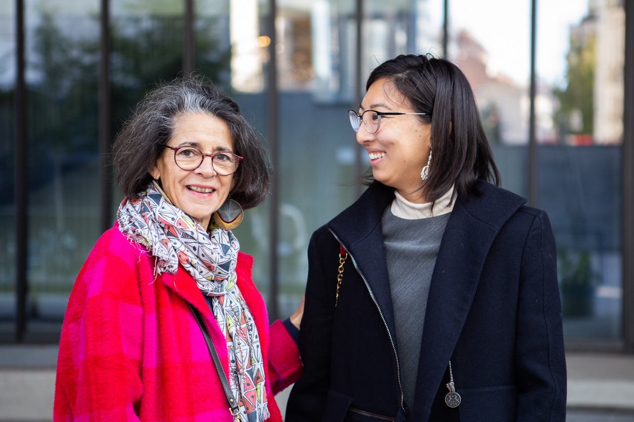 Un groupe de 4 personnes pose sur la terrasse d'un café, de droite à gauche, il y a un jeune homme avec des nattes assis sur le dos d'une chaise, un homme blanc de plus de 50 ans avec un pull rose qui s'appuie sur le premier. Ensuite une dame coupe courte et pull bleu dont l'épaule touche l'épaule de la dernière jeune femme à la peau foncée et de longues tresses