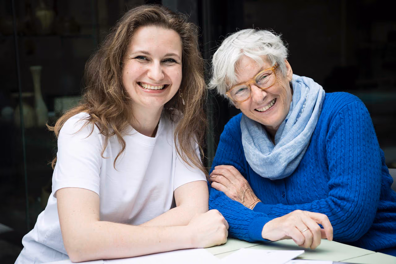 A young woman with long brown hair is smiling with all her teeth with her mentor next to her who is also smiling. The mentor is an elderly woman with white hair and glasses.