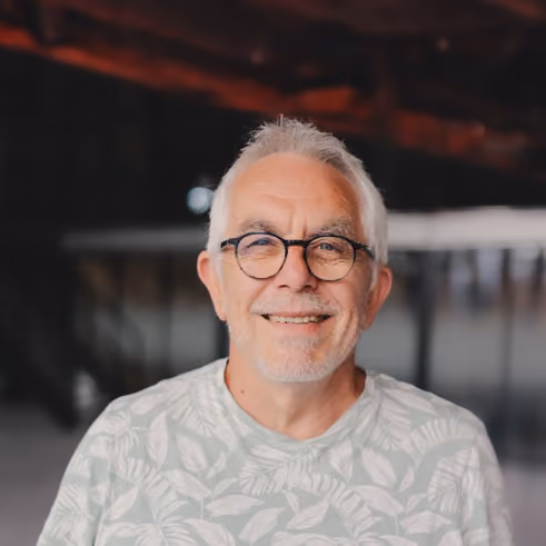 Portrait d'un homme avec les cheveux courts et blancs et des lunettes noires sourit en regardant le photographe. Il porte un tshirt vert pâle à motif feuilles