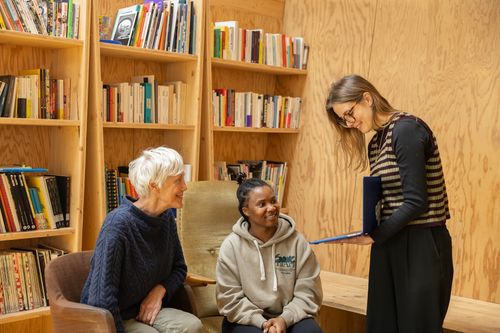 trois personnes regardent un écran d'ordinateur en souriant, un jeune homme d'origine arabe assis, une jeune femme debout avec des cheveux blonds et un homme plus âgé assis également