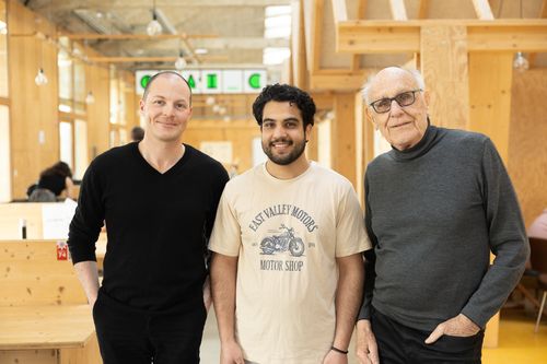 deux personnes sont assis à une table et discutent. La première est un homme de plus de 60 ans avec un pull couleur saumon, la seconde un homme noir avec un panama beige