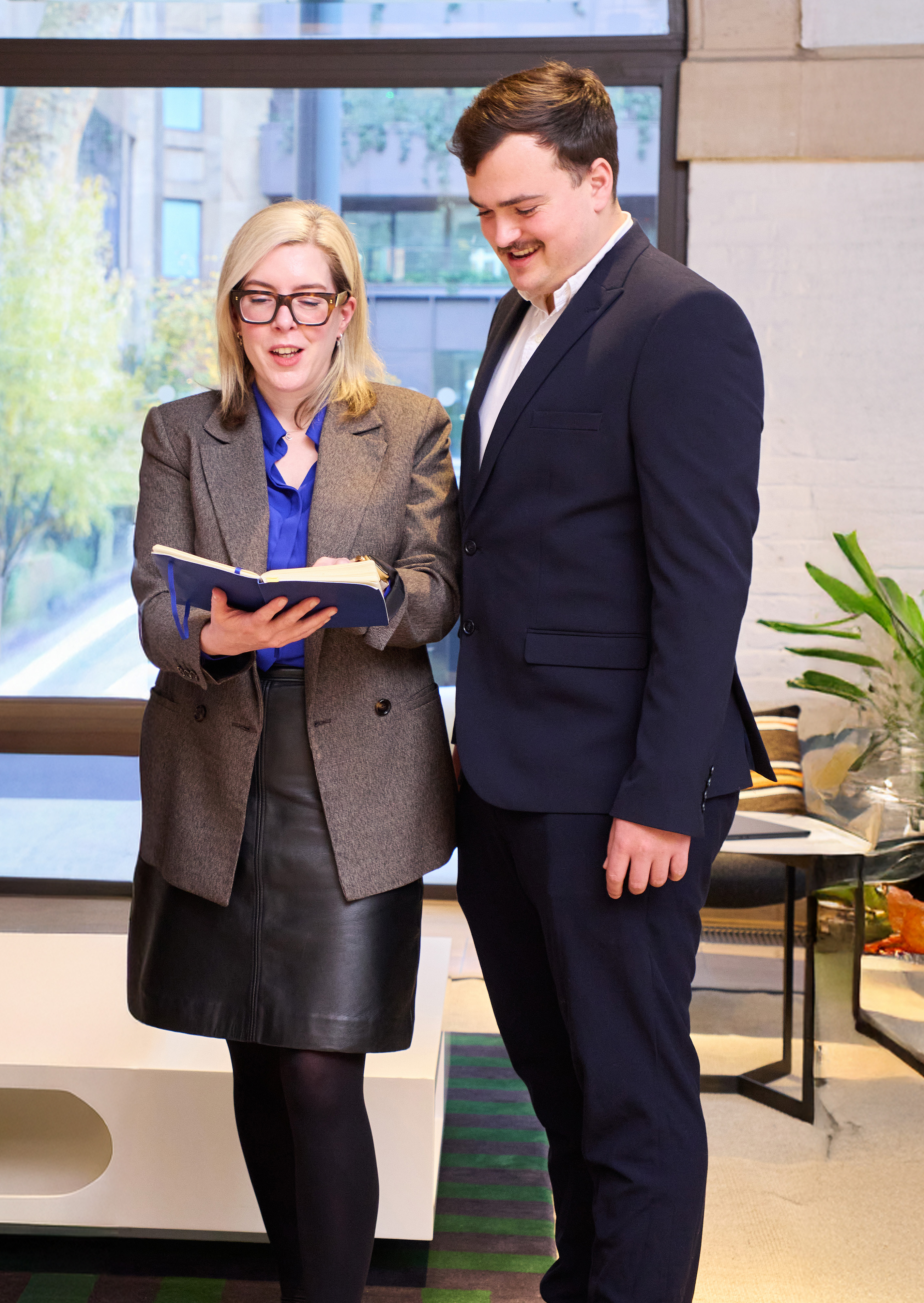 Two professionals standing indoors near a large window; she reads a notebook while a man in a dark suit looks on and smiles.