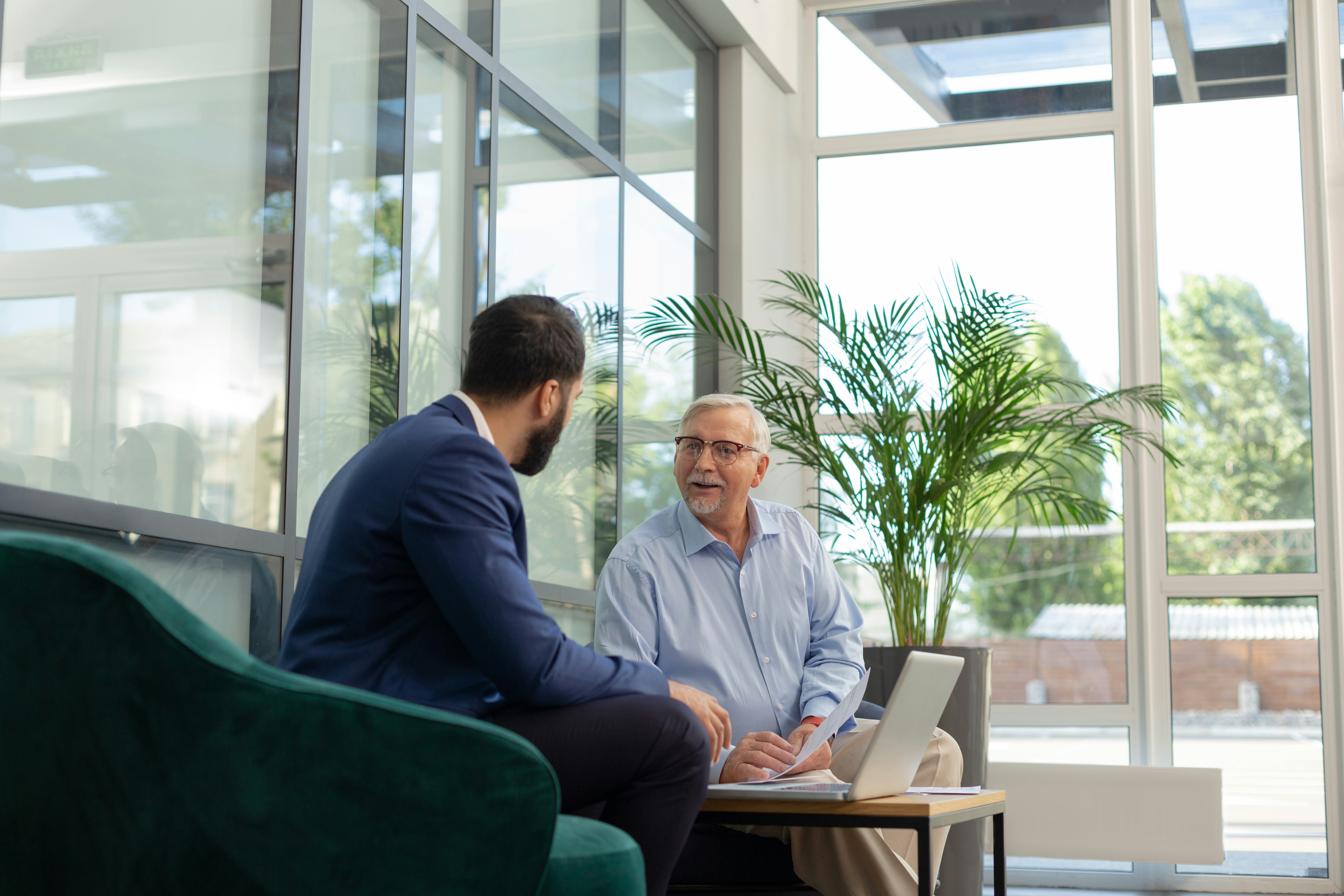 Two men having a discussion in a bright office, one holding papers and a laptop on a table in front of them.
