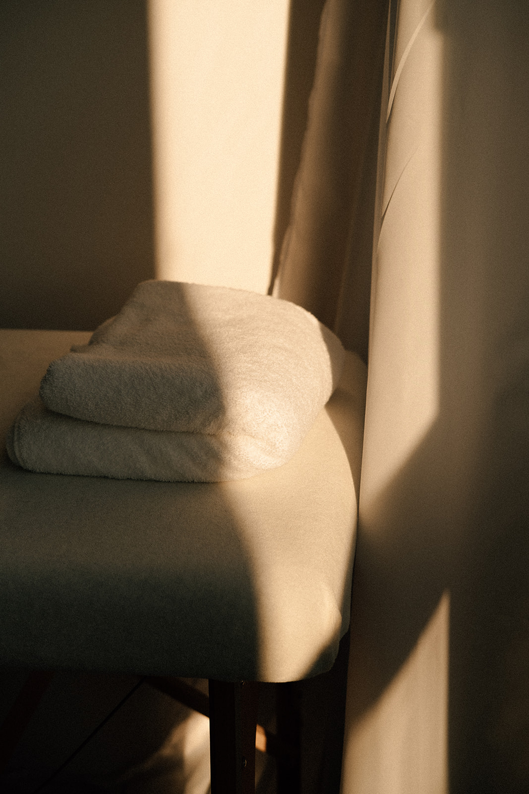 Eine Massageliege mit einem zusammengefalteten Frotteetuch. Ein warmer Sonnenstrahl strahlt über die Liege und das Tuch. 
