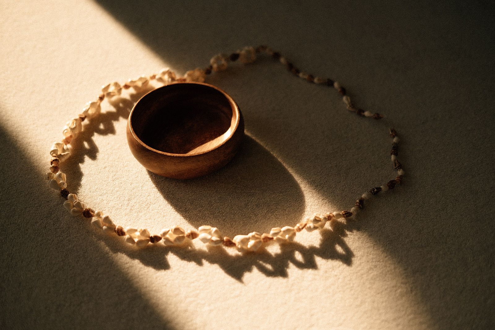 Eine leere Holzschale befindet sich auf einer Massageliege. Um die Schale befindet sich eine Muschelkette. Ein warmer Sonnenstrahl scheint von links oben auf die Schale und die Kette.