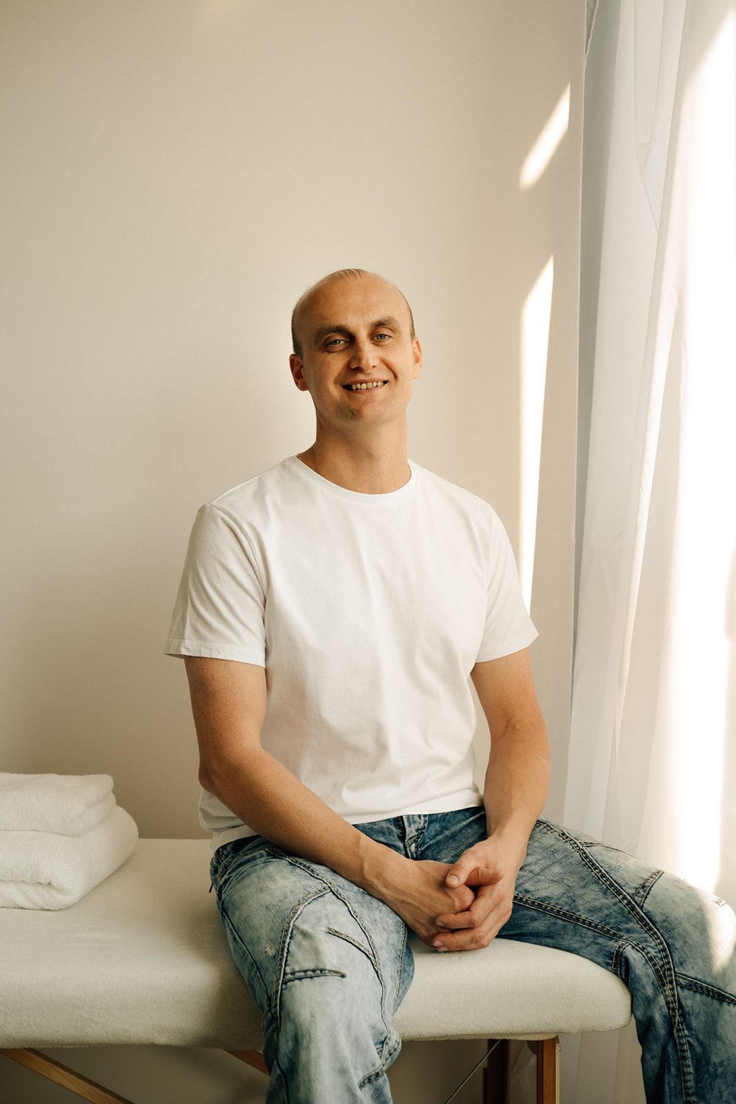 Ein Mann sitzt auf einer Massageliege. Er lächelt und sieht freundlich aus. Er trägt ein weisses T-Shirt und eine blaue Jeans.